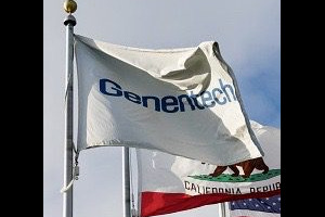 A photo of the Genentech flag waving in the wind, with the Genentech Campus buildings visible in the background beneath a clear blue sky.